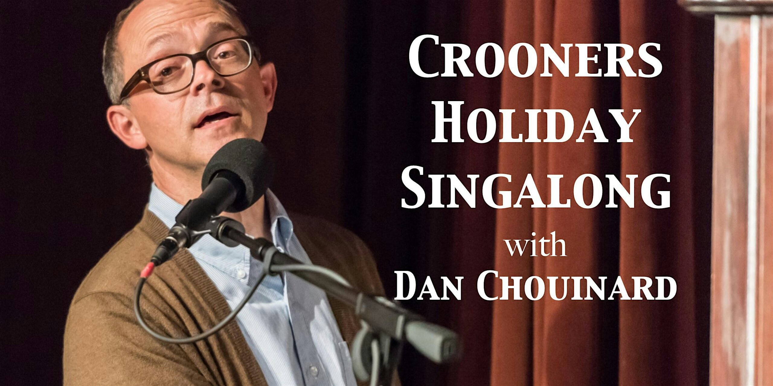 Man wearing glasses speaks into a microphone on stage; poster text on the right reads 'Crooners Holiday Singalong with Dan Chounard'.