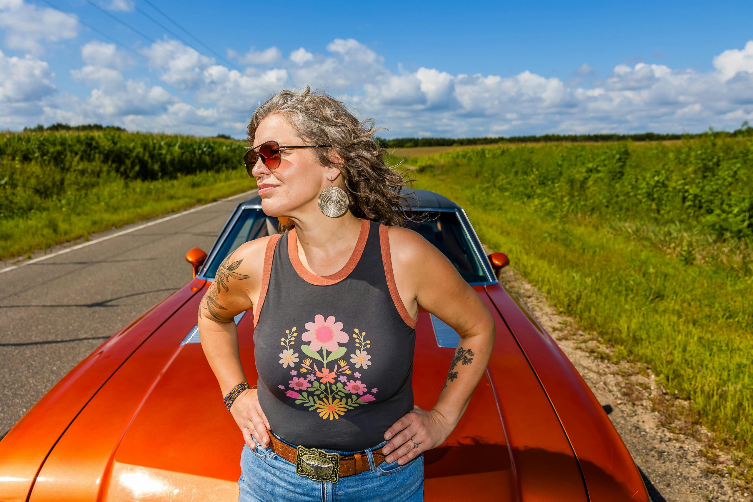 Woman wearing sunglasses and a floral tank top leans on an orange car along a rural road on a sunny day.
