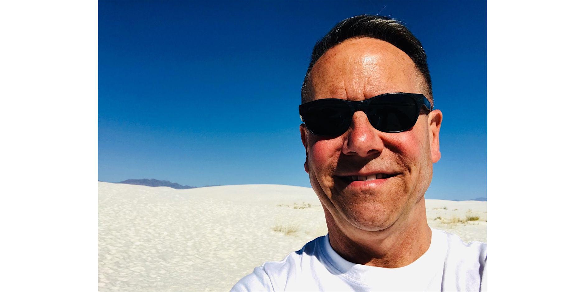 Close-up of a man wearing black sunglasses in a sunny desert setting with white dunes and a blue sky.