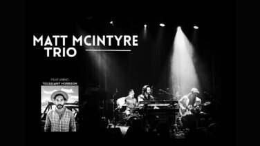 Black-and-white stage photo of the Matt McIntyre Trio performing under spotlights, with large 'MATT MCINTYRE TRIO' text on the left and a smaller portrait of Toussaint Morrison on the bottom left.