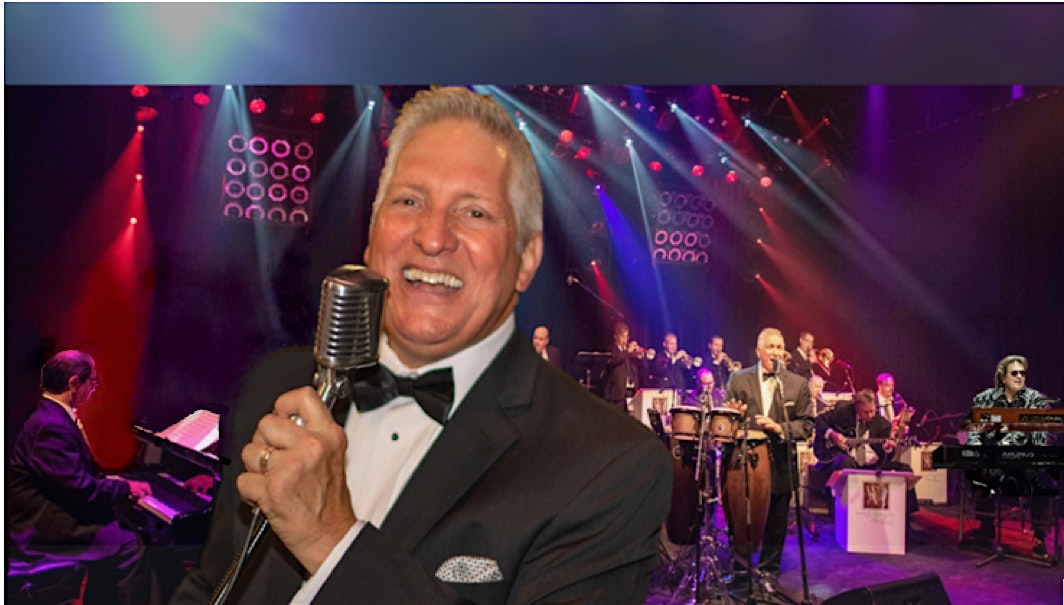 Smiling male singer in a tuxedo sings into a vintage microphone on stage with a full band behind him.