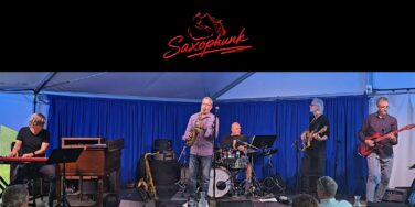 Live jazz band Saxophunk performing on a blue-curtained stage under a tent; keyboardist left, saxophonist center, drummer, bassist, and guitarist playing.
