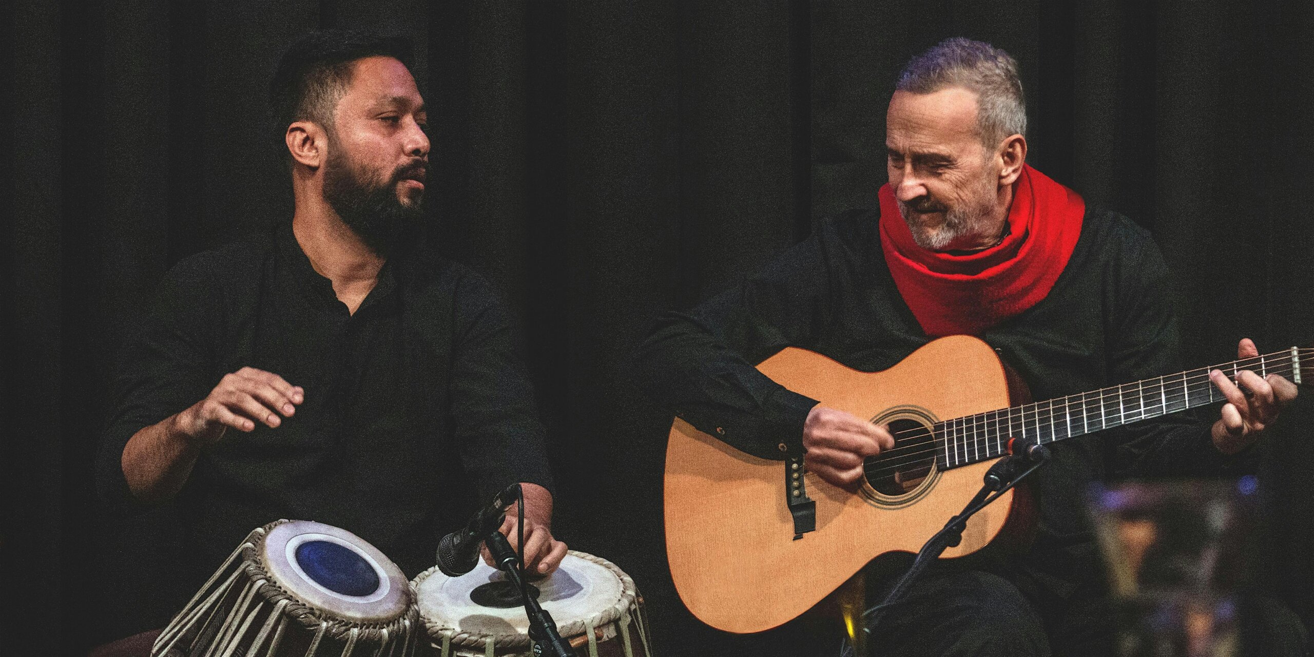 Two musicians on a dark stage: a guitarist in a red scarf strums an acoustic guitar while a percussionist plays hand drums beside him, both focused on music.