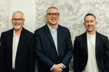 Three men in blazers posing for a group photo against a patterned wall; left with glasses and striped suit, center in a navy blazer, right in a black blazer and beige shirt.