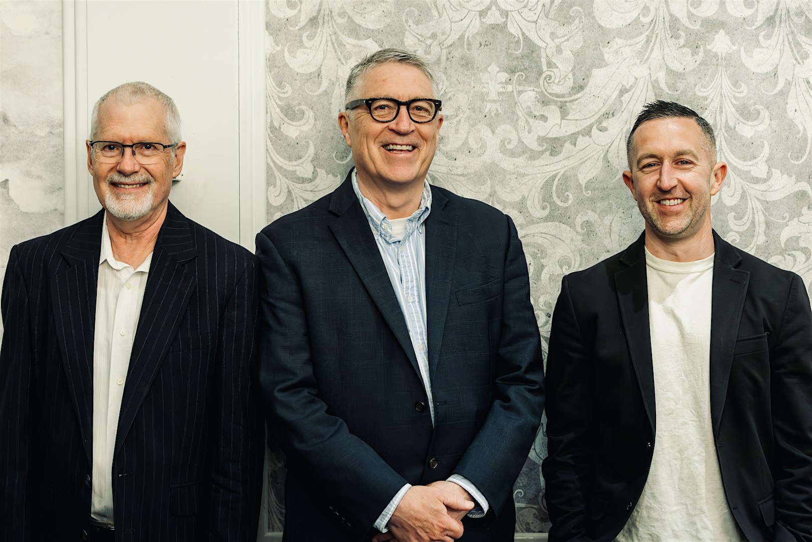 Three men in blazers posing for a group photo against a patterned wall; left with glasses and striped suit, center in a navy blazer, right in a black blazer and beige shirt.