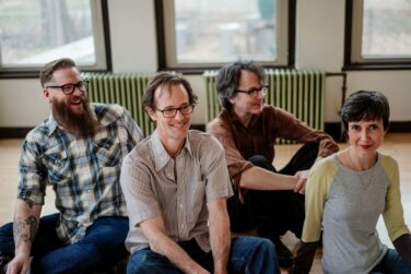 Four adults seated on the floor in a bright room, smiling with windows and radiators visible in the background.