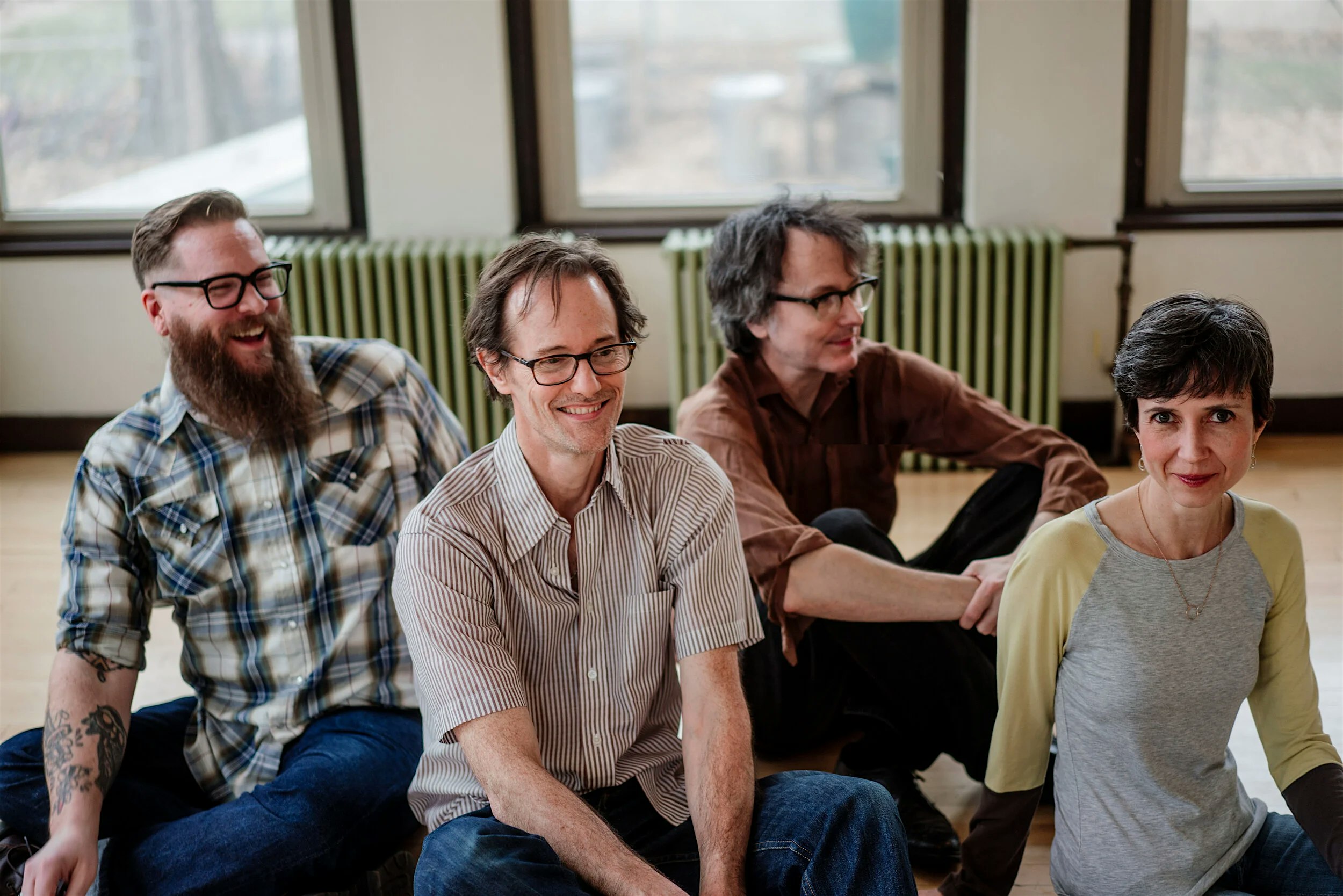 Four adults seated on the floor in a bright room, smiling with windows and radiators visible in the background.