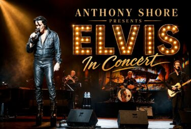 Elvis tribute concert on a dark stage with a lead singer in a black leather outfit, large illuminated ELVIS marquee lettering, and a full band in the background under warm spotlights.