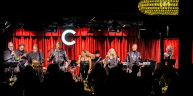 Live band performing on a red-curtained stage, with two female vocalists center and a full ensemble of musicians on either side, in front of a dark audience. The large illuminated C is visible in the background.
