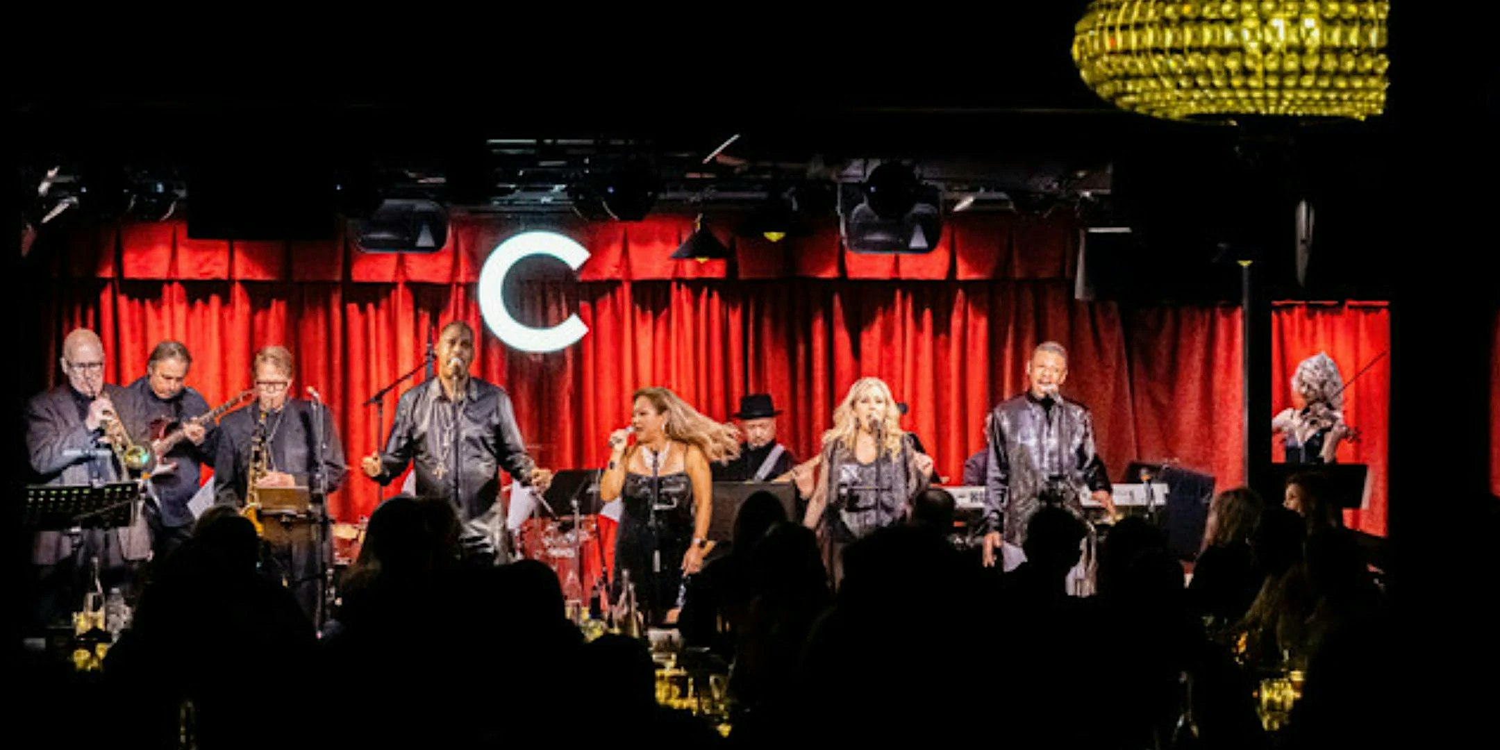 Live band performing on a red-curtained stage, with two female vocalists center and a full ensemble of musicians on either side, in front of a dark audience. The large illuminated C is visible in the background.