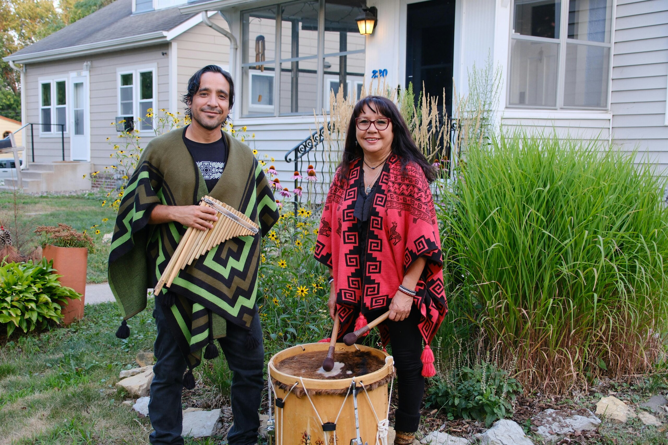 Two people stand in a garden in front of a house, wearing colorful ponchos and holding percussion instruments (drum and wind chime).