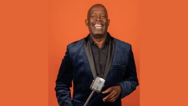 Smiling man in a blue velvet suit holding a retro microphone against an orange background.
