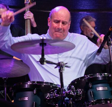 Drummer in a light blue shirt performing on stage with a drum set and purple lighting, mid-performance diverging sticks in motion.