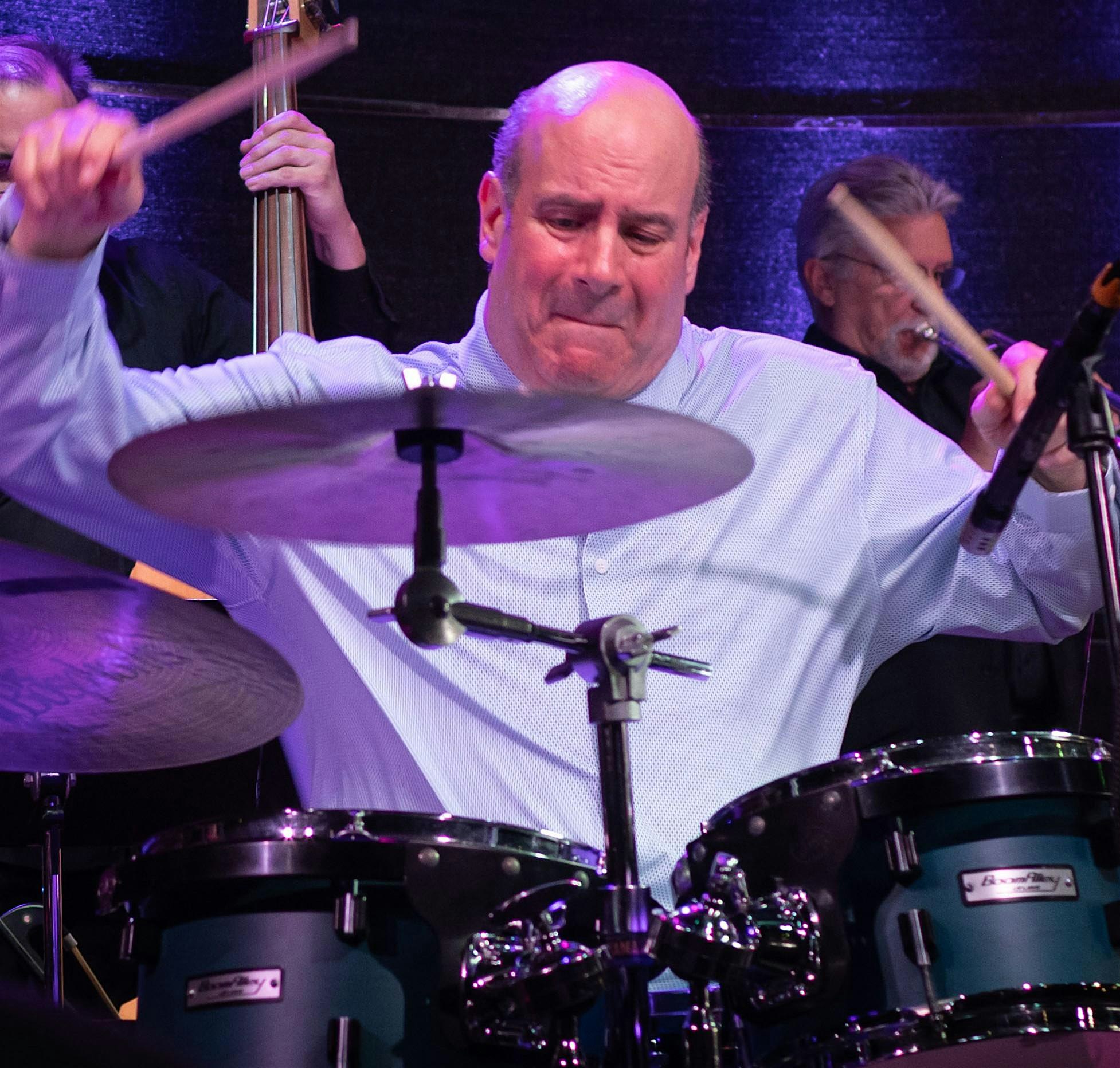 Drummer in a light blue shirt performing on stage with a drum set and purple lighting, mid-performance diverging sticks in motion.