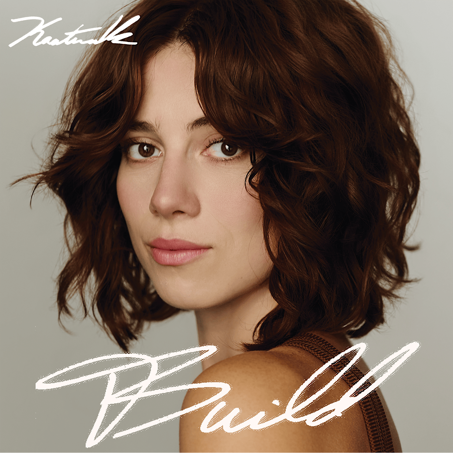 Close-up portrait of a woman with wavy brown hair, looking slightly to the left, with white cursive text overlaid on a gray background.