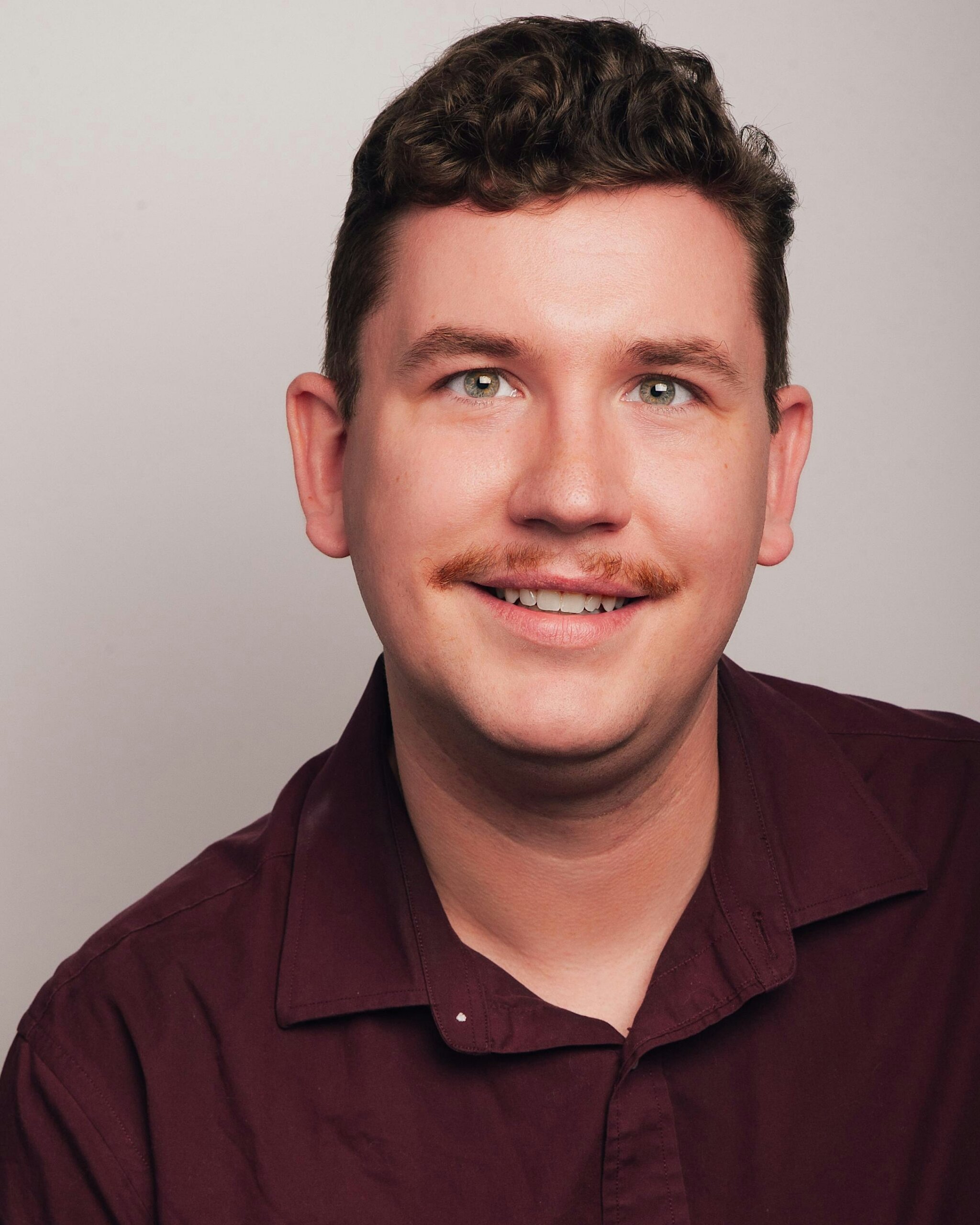 Close-up of a smiling man with short dark hair and a light beard, wearing a burgundy shirt against a neutral background.