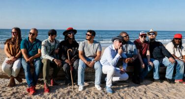 A diverse group of friends sits closely together on a wooden seawall on a sunny beach, with the ocean behind them.