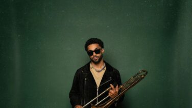 Male musician wearing sunglasses and a black jacket, holding a trombone against a green backdrop.