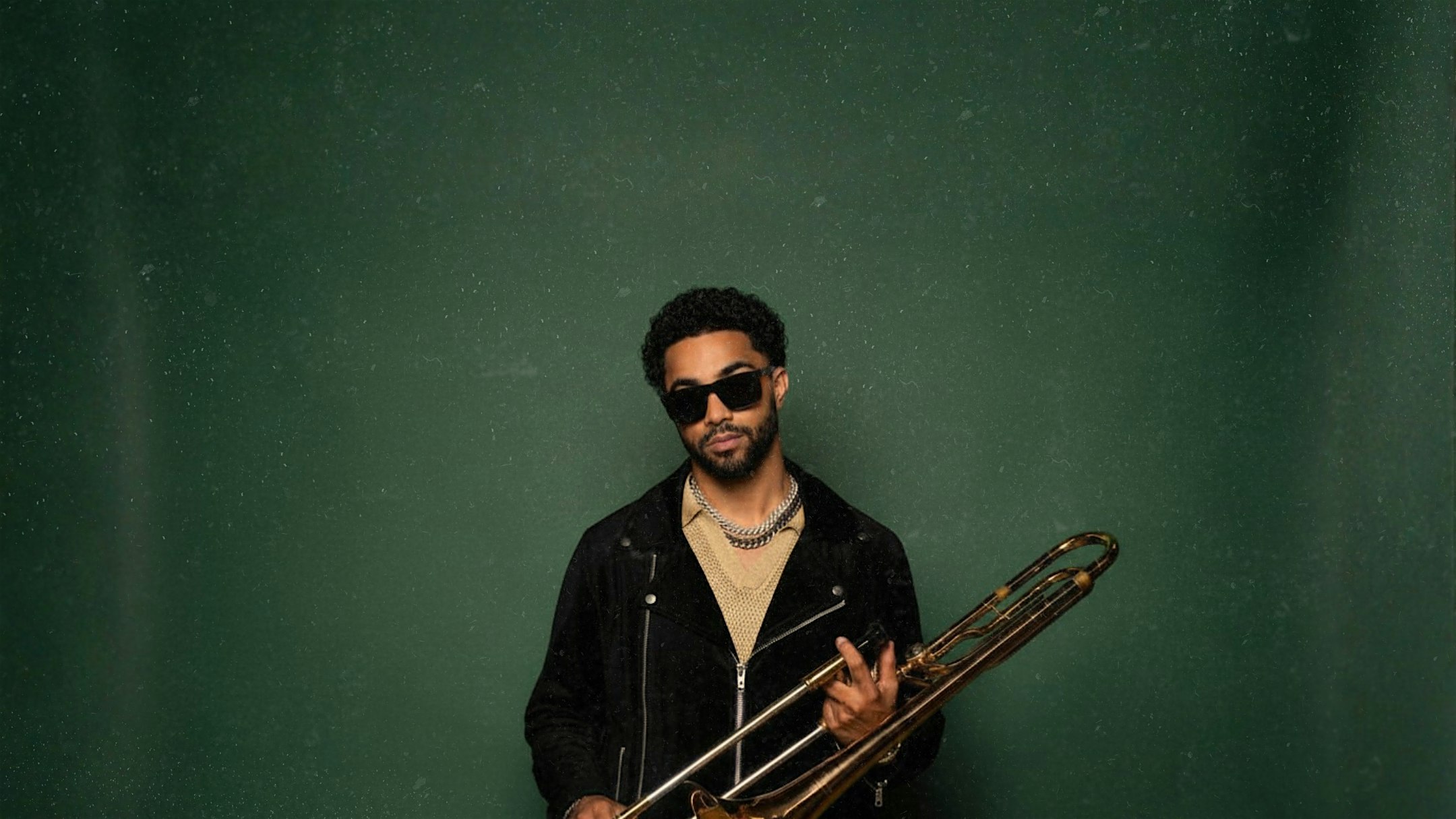 Male musician wearing sunglasses and a black jacket, holding a trombone against a green backdrop.