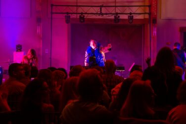 Live concert scene with a lead singer in a blue feather boa performing center stage under magenta lighting, audience in foreground.