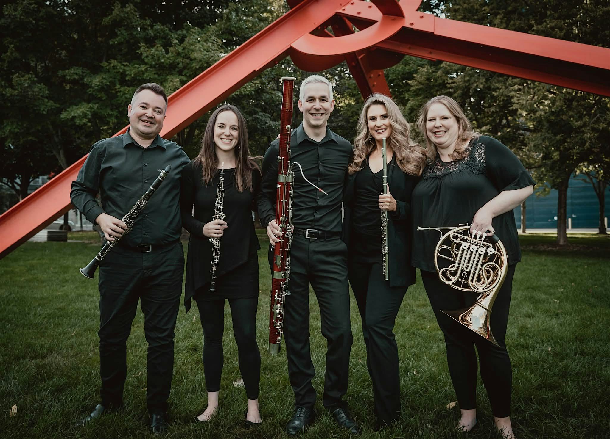 Five musicians in black, standing outdoors with their wind instruments, posing in front of a red metal sculpture and green grass.