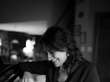 Woman with dark hair smiling, resting her hand on a piano keyboard in a softly lit room.