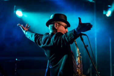 Male performer on a dark stage with blue lights, wearing a black hat and sunglasses, arms raised as he sings into a microphone with a saxophone nearby