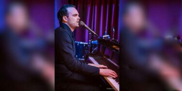 Male pianist singing into a microphone while playing a grand piano on a purple-lit stage with drums in the background