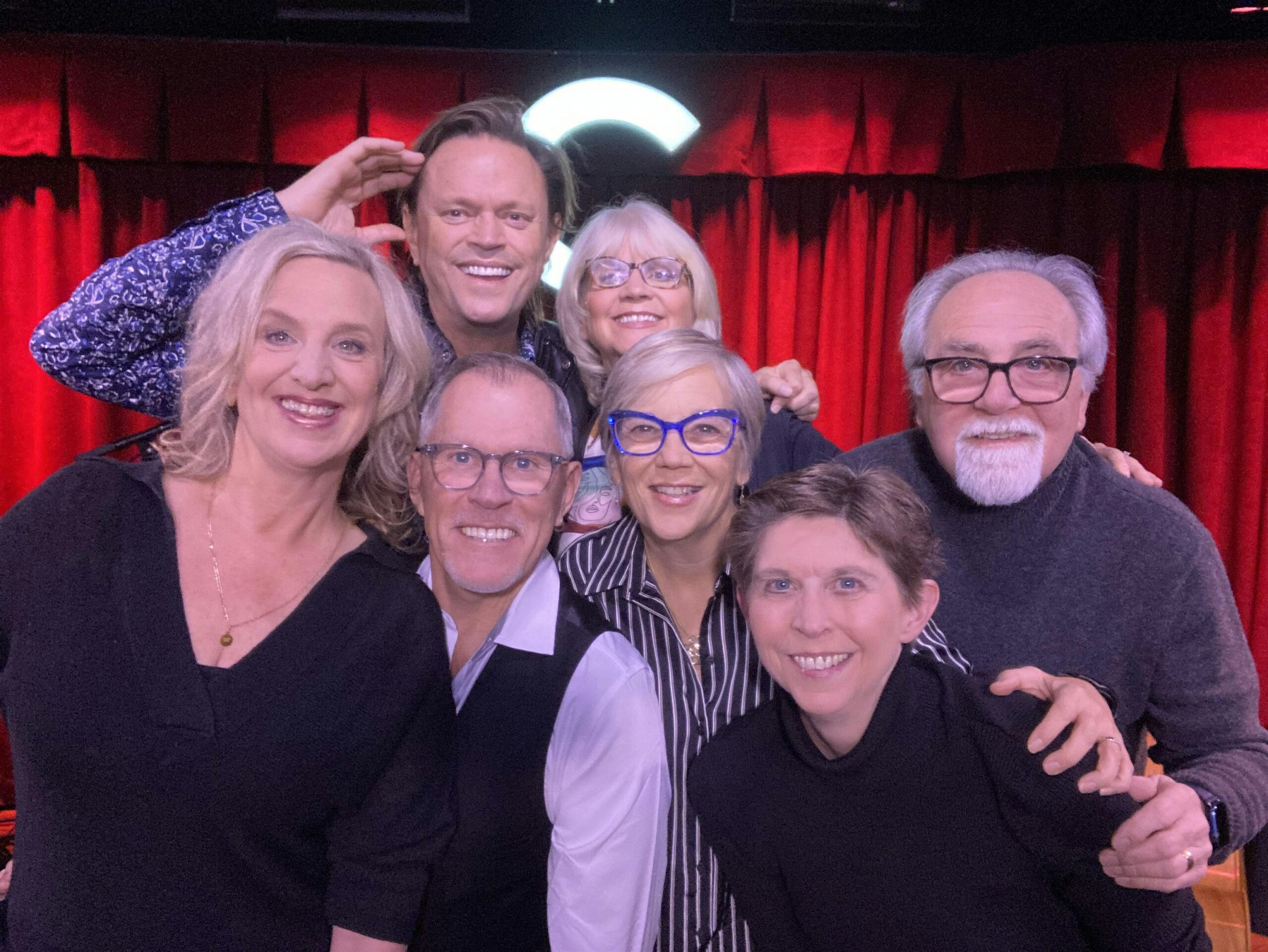 Group of smiling adults posing closely for a photo in front of red theater curtains on a stage.