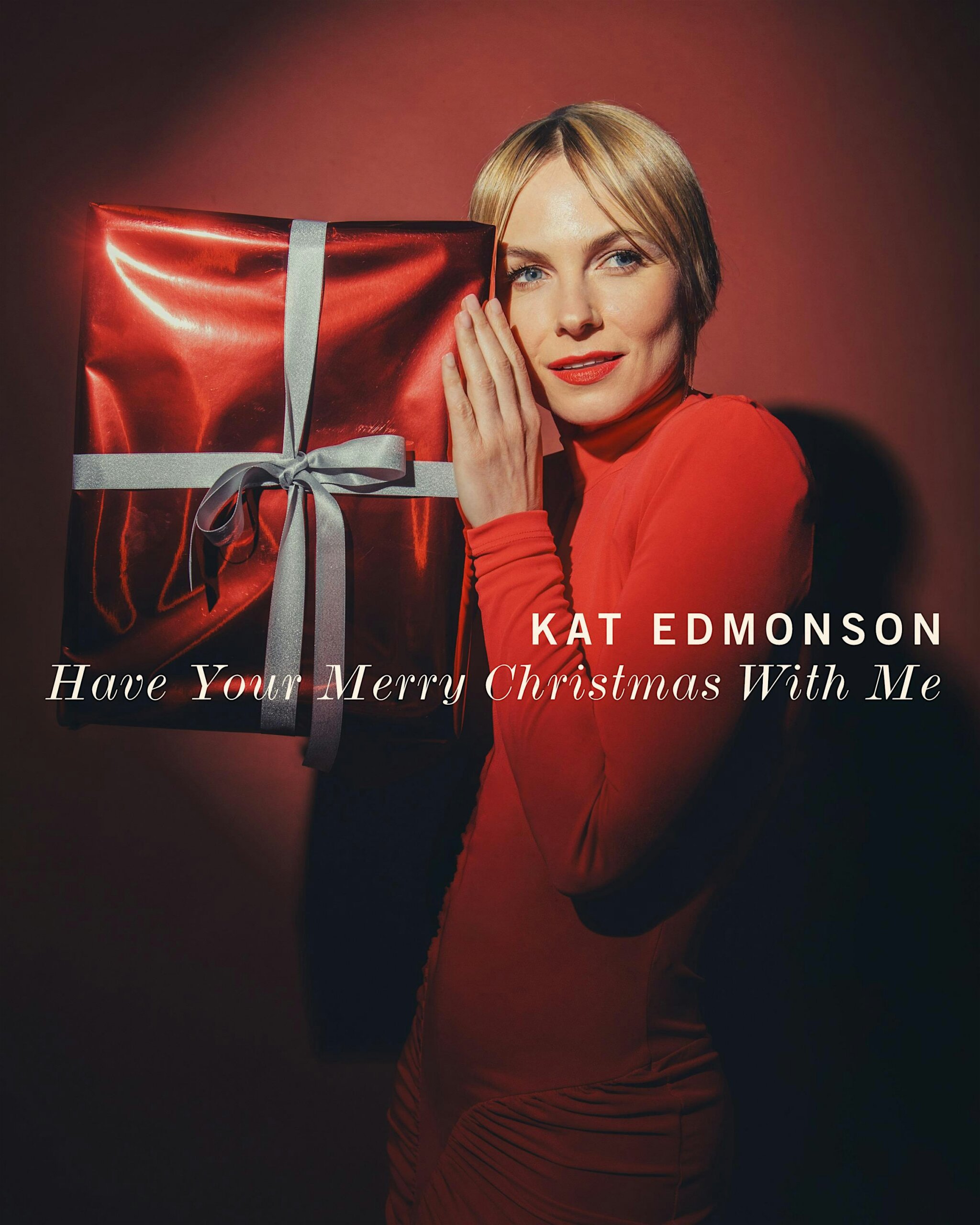 Woman in red holds a large red gift box with a silver ribbon, smiling at camera against a dark red backdrop (text reads 'Have Your Merry Christmas With Me' and 'Kat Edmonson').