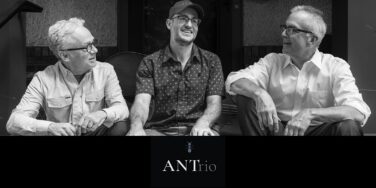 Black-and-white photo of three men sitting together outdoors, smiling, with a logo reading ANTriO below them.