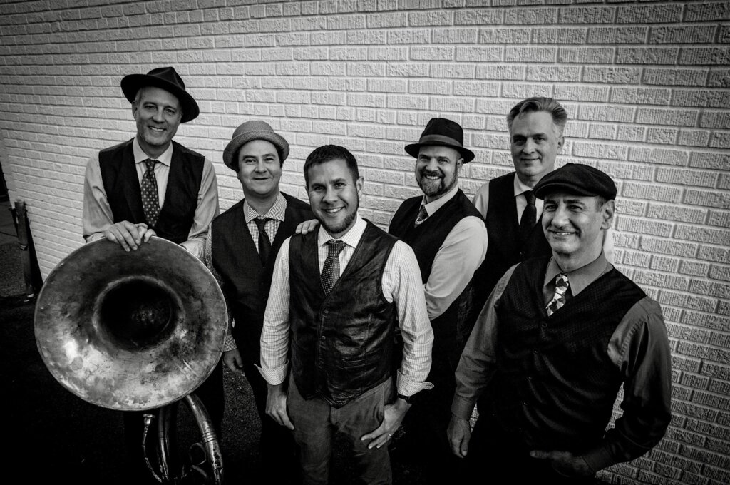 Six male musicians in vintage vests and hats pose with a large brass tuba against a brick wall nearby.