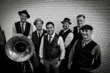 Six male musicians in vintage vests and hats pose with a large brass tuba against a brick wall nearby.