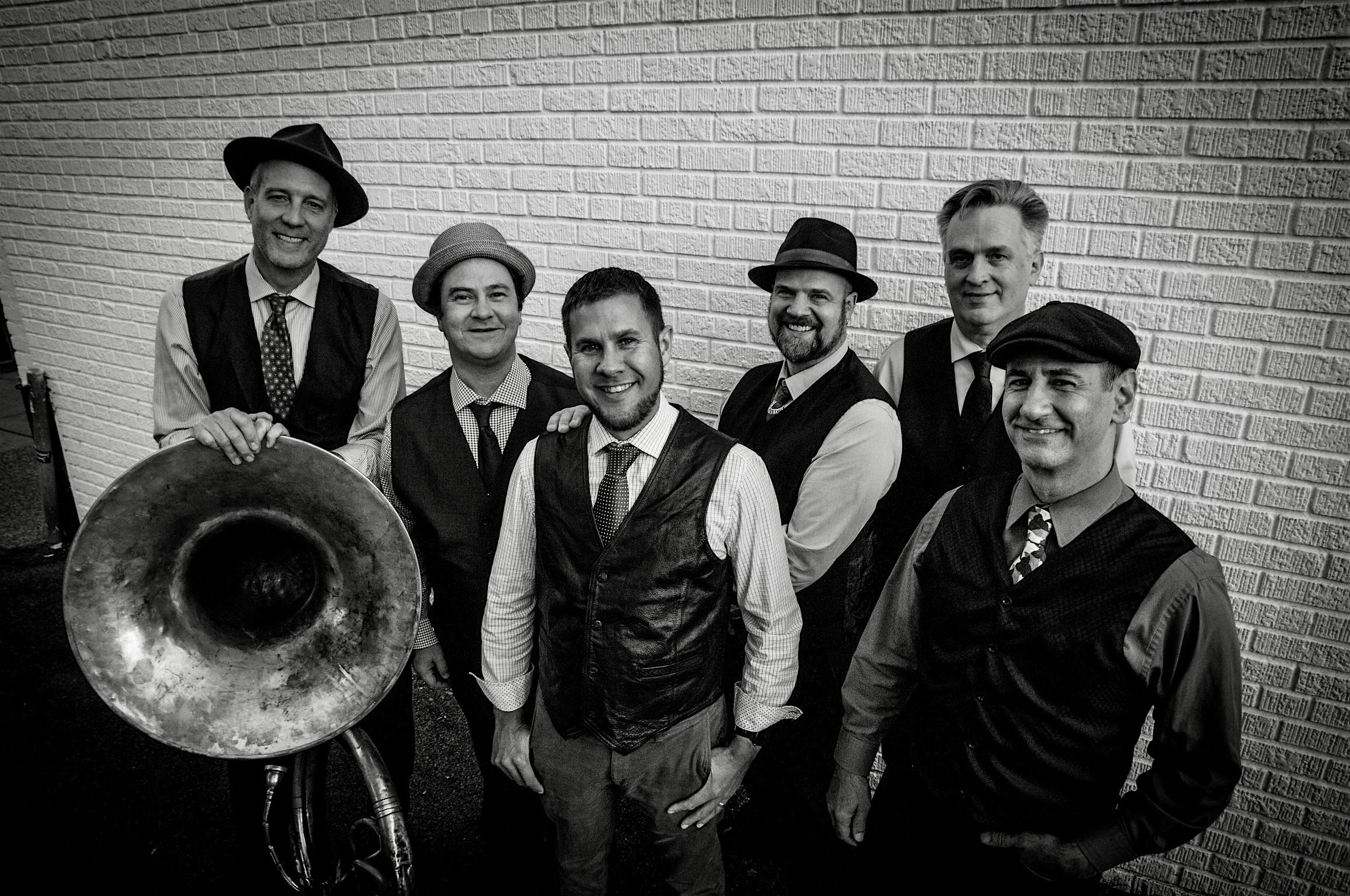 Six male musicians in vintage vests and hats pose with a large brass tuba against a brick wall nearby.