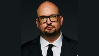 Man with a shaved head and black-framed glasses in a dark suit and white shirt, facing the camera against a dark background.