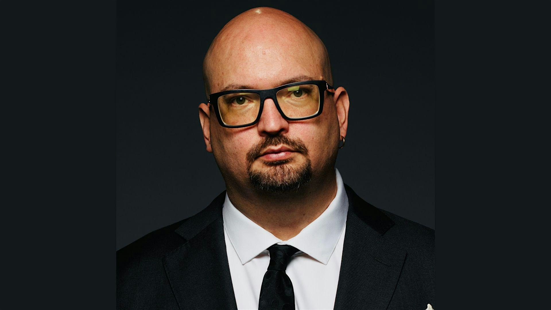 Man with a shaved head and black-framed glasses in a dark suit and white shirt, facing the camera against a dark background.