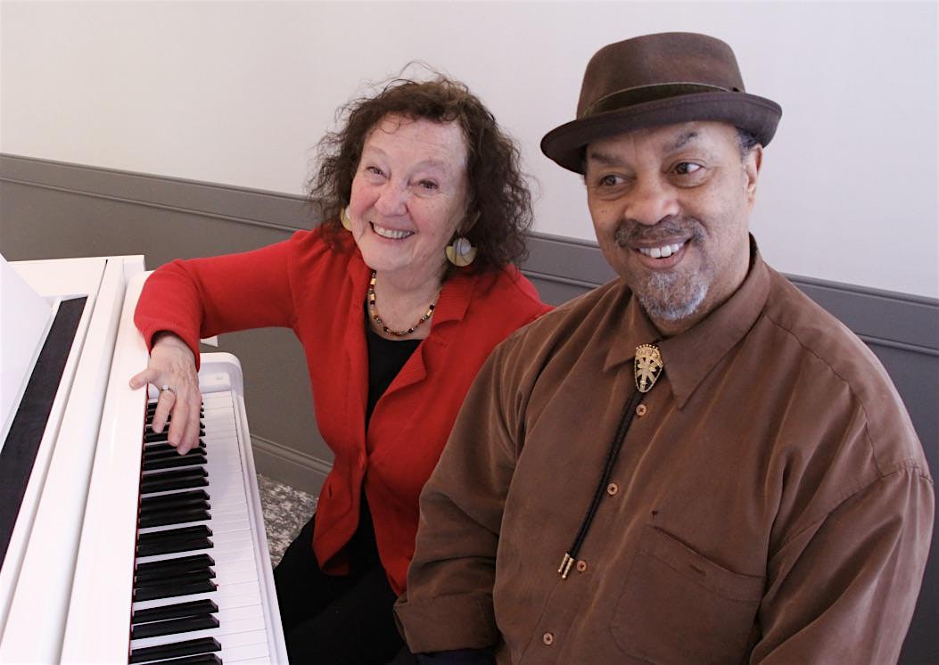 Two smiling adults sit at a white piano, the woman in red and the man in a brown shirt wearing a hat pose for the photo.
