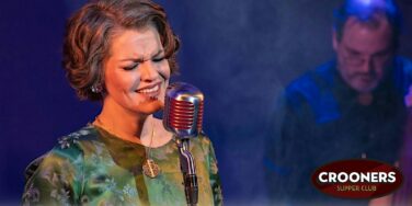 Female singer in a green floral dress sings into a vintage silver microphone on stage, Crooners Supper Club logo in the corner.