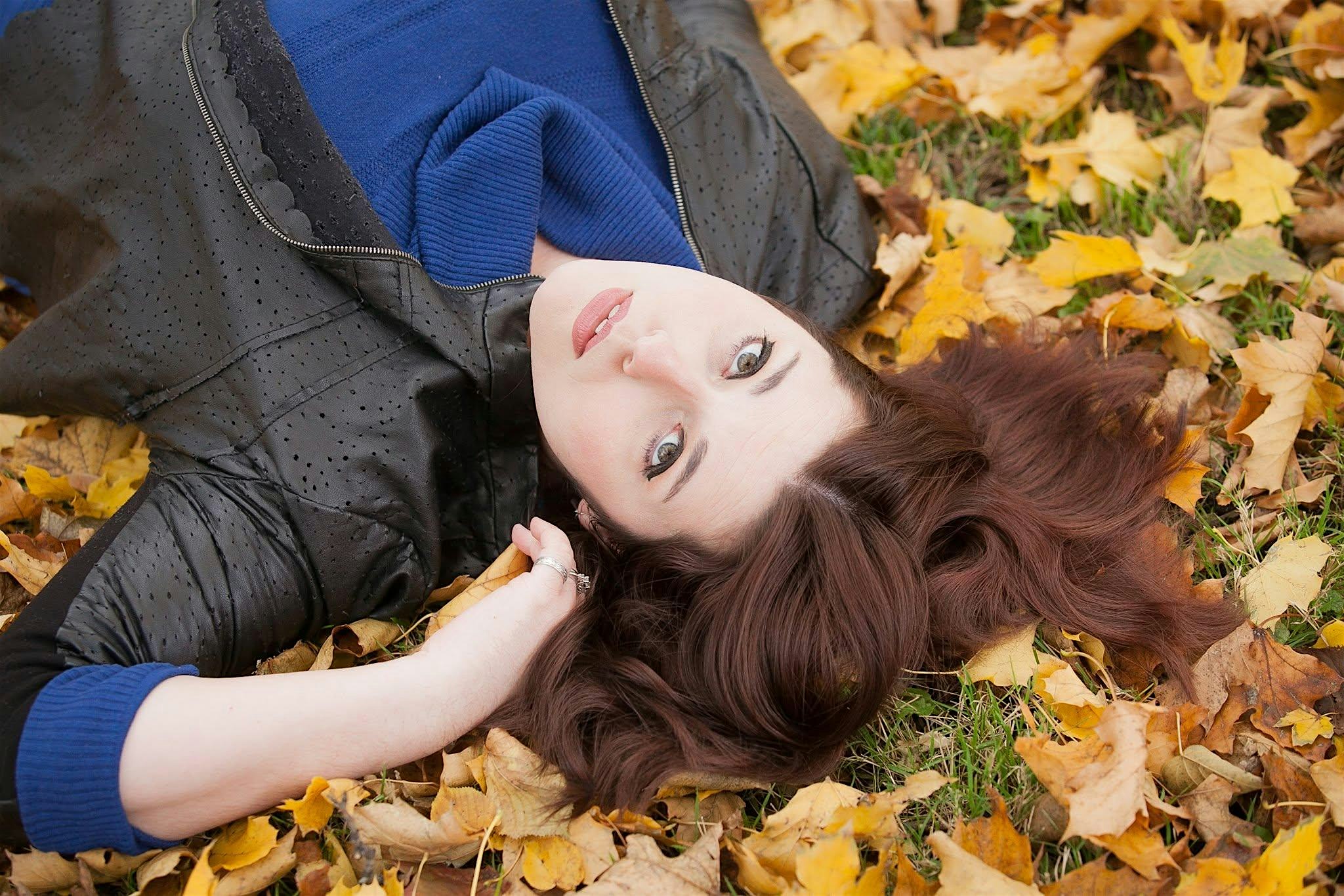 Woman lying on autumn leaves, wearing a blue sweater and a black dotted jacket.