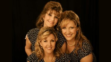 Three women in black-and-white polka-dot dresses pose together, smiling against a dark backdrop.