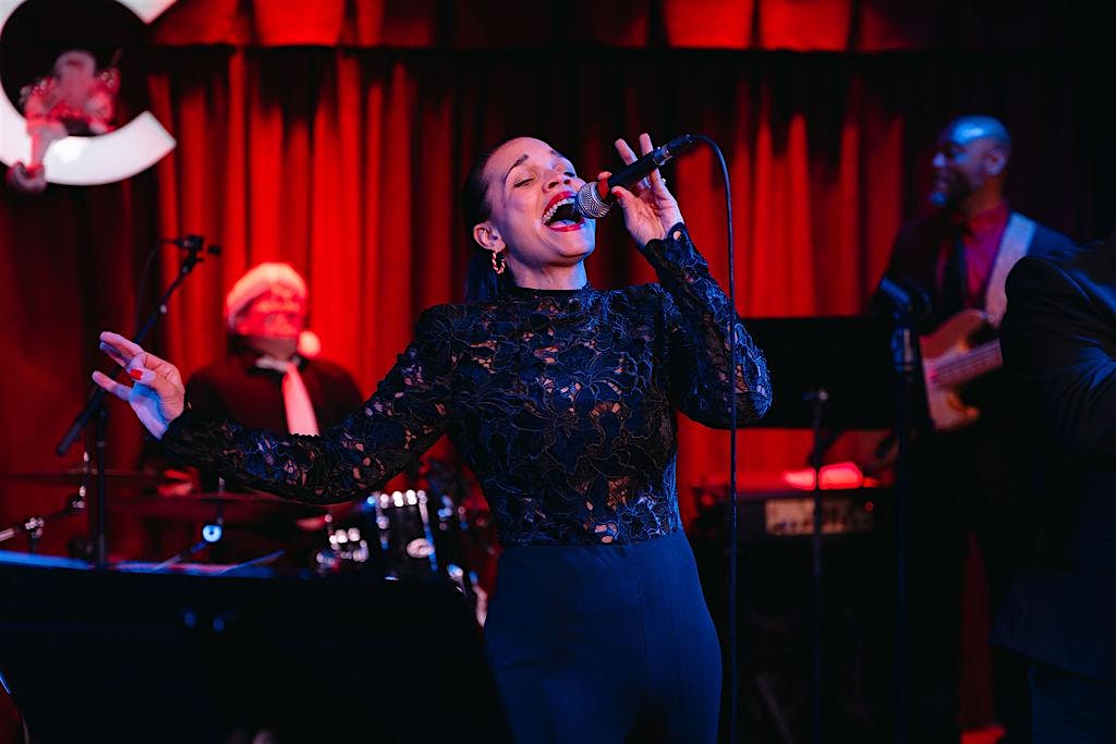 Female vocalist in a black lace top sings into a microphone on a red-lit stage with a live band in the background (drums and bass).