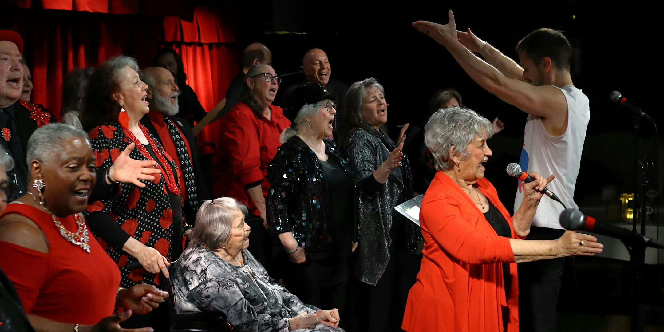 A diverse group of seniors singing on a stage with red curtains, many in red or patterned outfits, reaching toward the audience.
