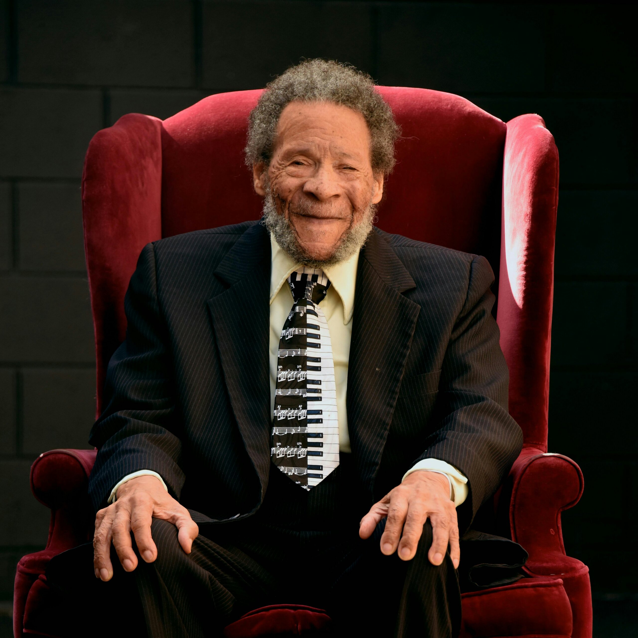 Elderly man with a gray beard sits in a red chair, smiling warmly at the camera in a dark room.