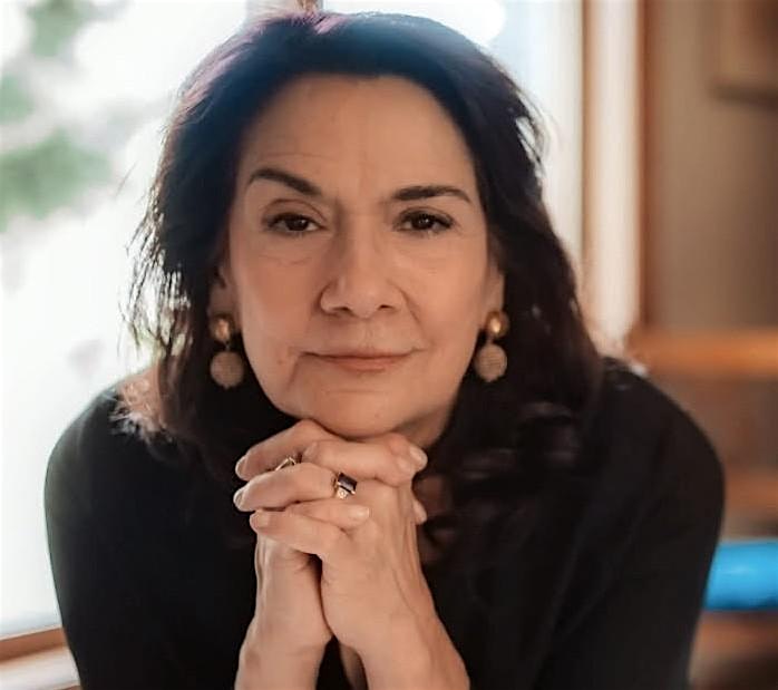 Portrait of a mature woman with dark hair, resting her chin on clasped hands, wearing gold earrings indoors near a window, looking thoughtfully at the camera.