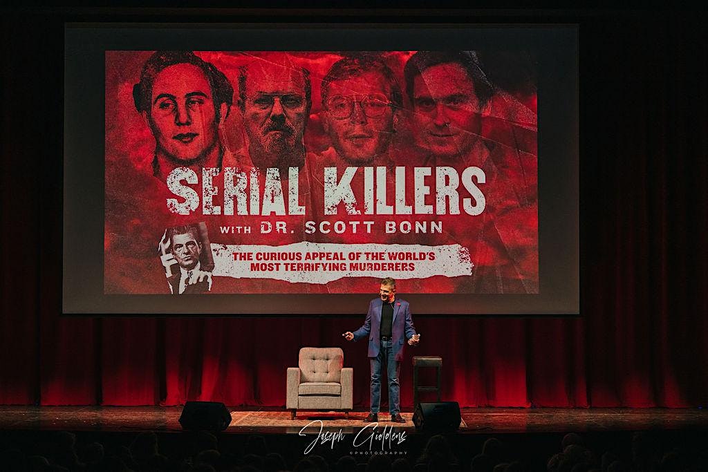 Man on stage presenting beside a red promotional poster for 'Serial Killers with Dr. Scott Bonn' on a large screen behind him.
