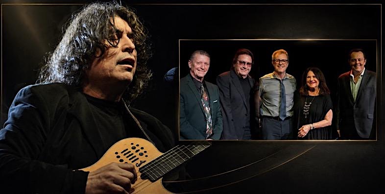 Guitarist with a curved acoustic guitar on stage, while a framed group photo of five people appears on the dark backdrop behind him.