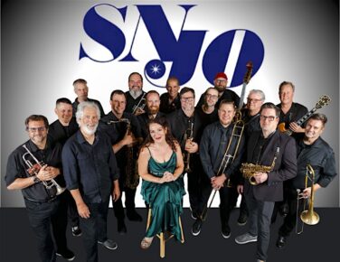 Large jazz band group photo: about 15 musicians in black, with a woman in a green dress seated center, surrounded by trumpets, trombones, and saxophones; blue logo backdrop.