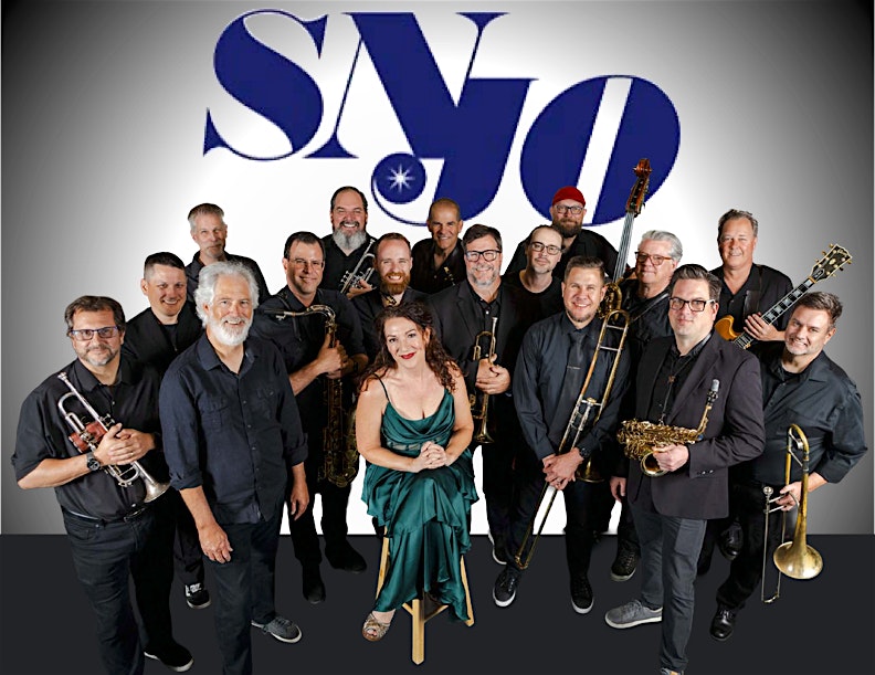 Large jazz band group photo: about 15 musicians in black, with a woman in a green dress seated center, surrounded by trumpets, trombones, and saxophones; blue logo backdrop.