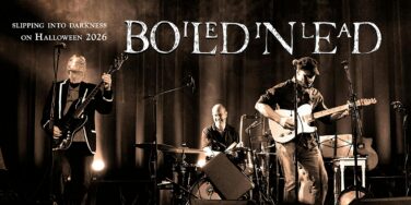 Live rock band on stage performing with guitars and drums under a dark backdrop; banner reads 'Slipping into Darkness on Halloween 2026' in the corner.