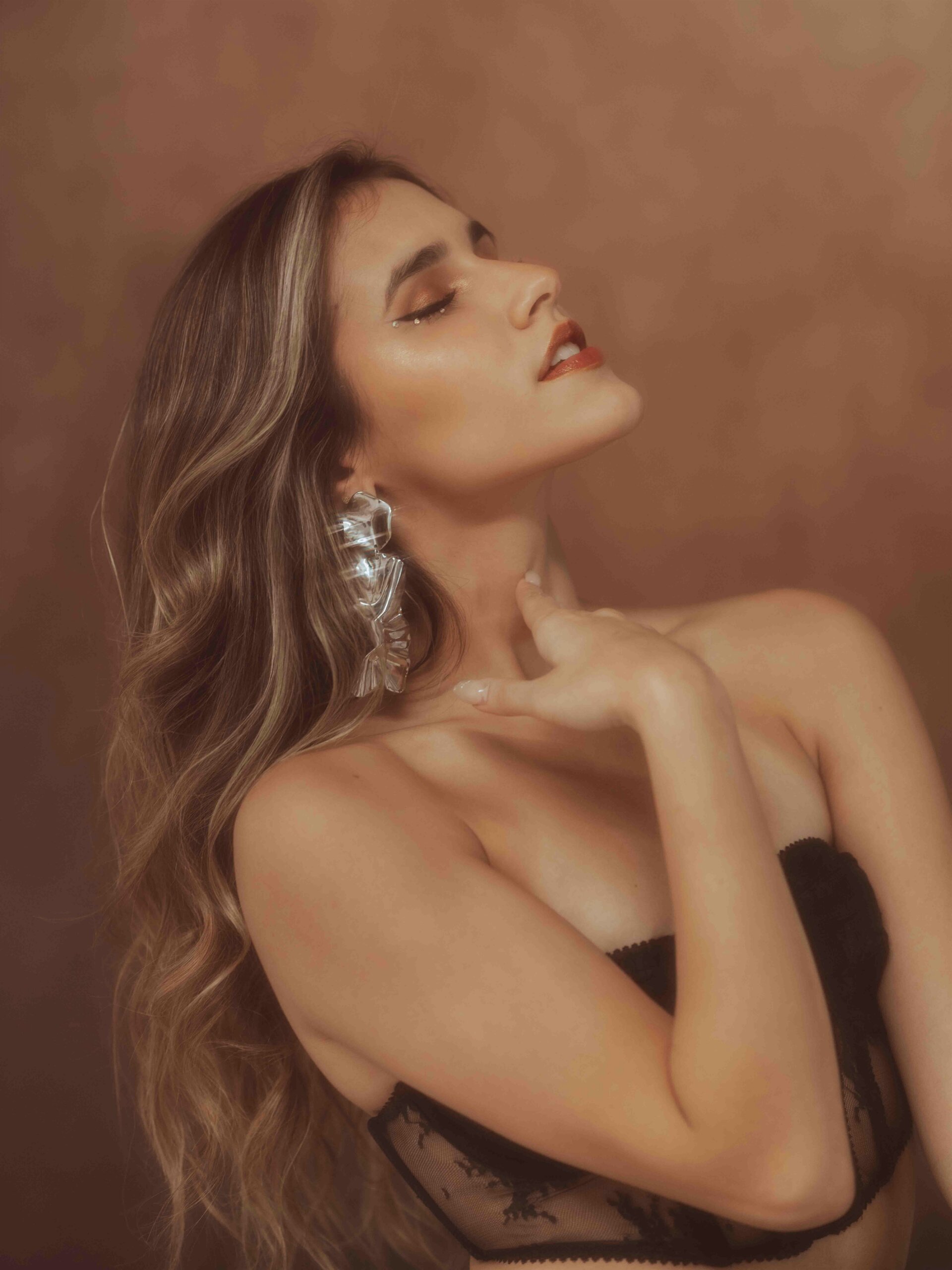 Woman with long wavy hair wearing black lace lingerie, head tilted back, eyes closed, touching her neck.